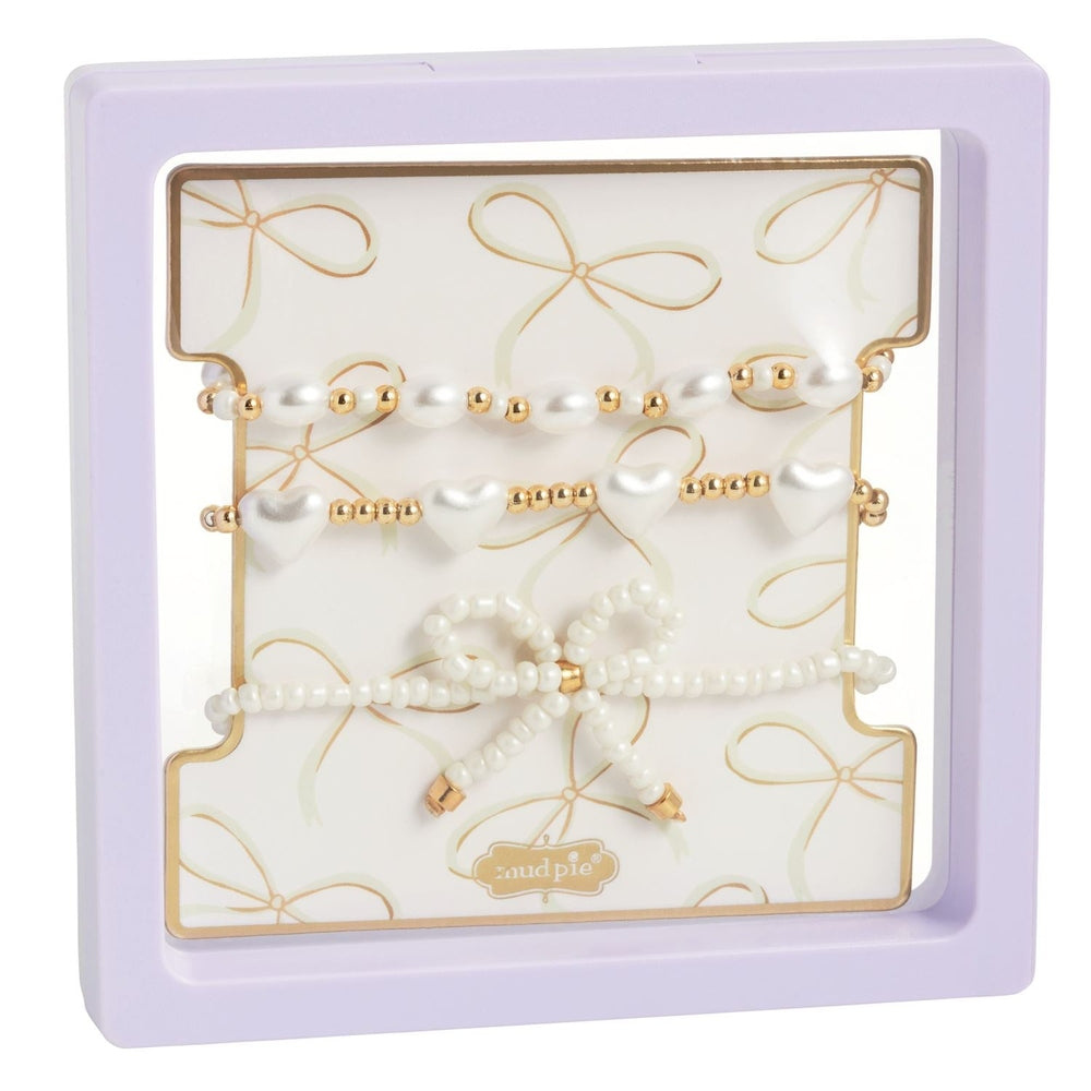 Beaded jewelry set by Mud Pie featuring three white and gold bracelets with heart-shaped charms and a central beaded bow displayed in a lavender square frame on a gold bow-patterned backing card