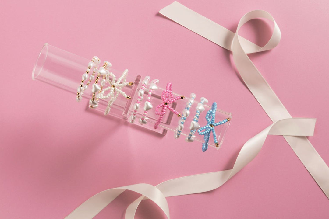 Studio photograph of handmade beaded jewelry displayed on a clear acrylic stand against a pastel pink background featuring heart-shaped pearls, pink and light blue beaded bows, and a winding white satin ribbon