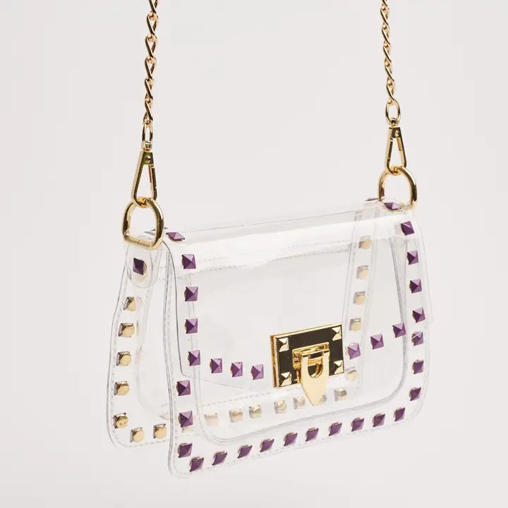Photograph of a transparent PVC handbag featuring purple and gold-toned pyramid studs and a gold turn-lock clasp against a neutral white background