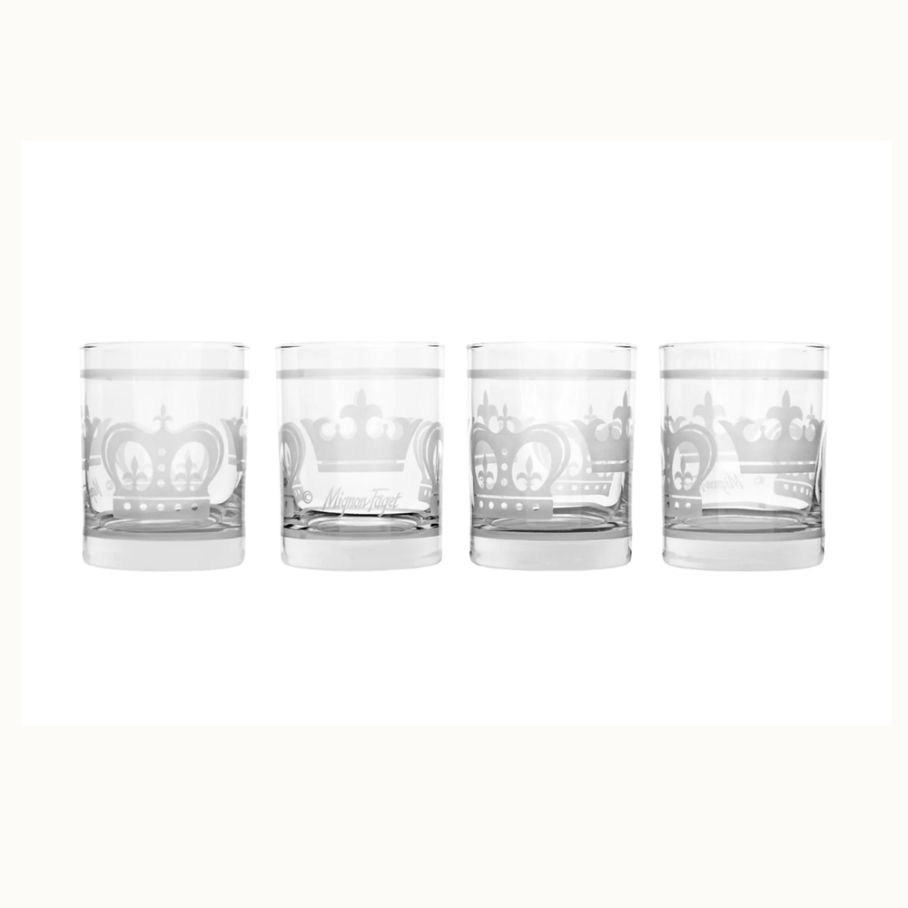 Etched glass by Mignon Faget featuring a set of four rocks glasses with frosted Mardi Gras crown motifs, fleur-de-lis accents, and heavy weighted bases on a white background