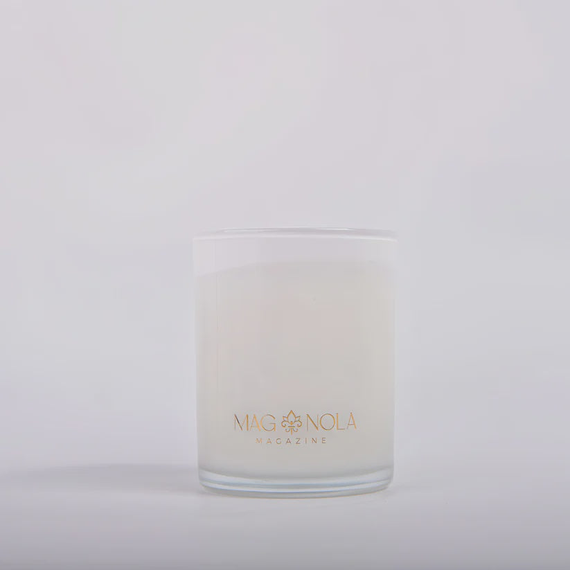 Photograph by Magnolia Magazine of a white scented candle in a frosted glass jar featuring a gold-toned fleur-de-lis emblem and elegant branding against a minimalist studio background