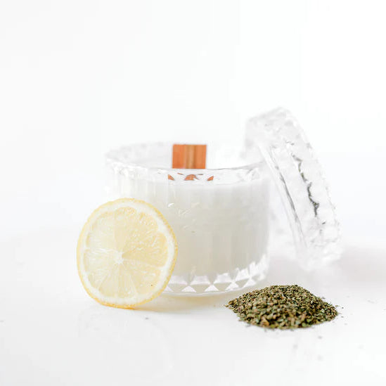 Photograph of a white scented candle in a diamond-cut crystal jar with a wooden wick featuring a vibrant yellow lemon slice and a small heap of dried green herbs on a minimalist white surface