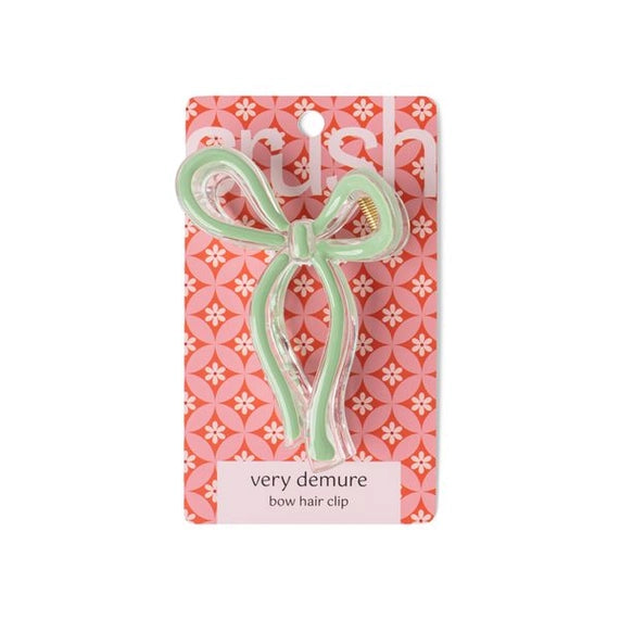 Commercial accessory by Crush, this mint green bow-shaped hair clip is presented on a pink floral patterned card featuring clear acrylic edges, a gold metal spring, and the label very demure bow hair clip