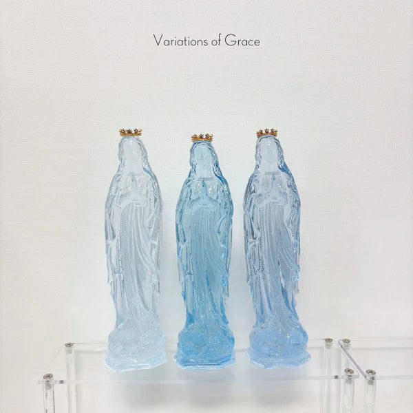 Translucent resin sculptures from the Variations of Grace collection featuring three iconic Virgin Mary figures in shades of cerulean and sky blue with miniature gold crowns and praying hands atop a clear acrylic display shelf