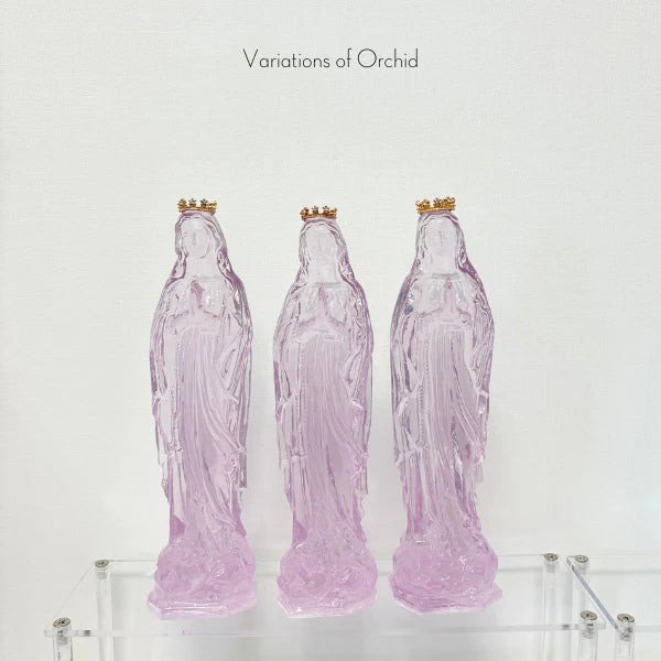 Translucent resin sculptures from the Variations of Orchid series depicting three Virgin Mary figurines on a clear acrylic shelf, highlighting their soft lavender-pink transparency and delicate gold crowns with tiny jewel embellishments