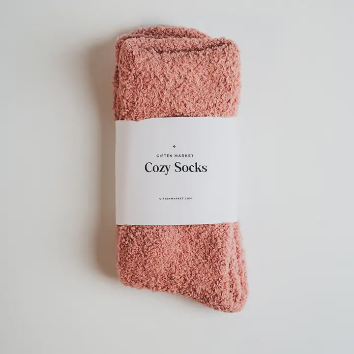 Product photograph by Giften Market of a pair of dusty rose fuzzy socks wrapped in a white paper band labeled Cozy Socks featuring black serif typography and a soft plush chenille texture against a minimalist white background