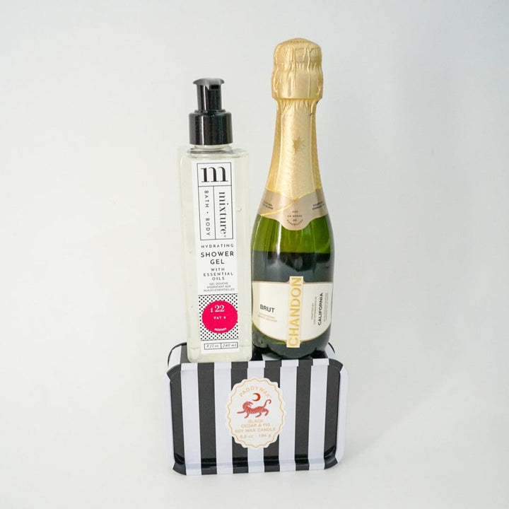 Studio photograph of a luxury self-care gift set featuring a bottle of Mixture Hydrating Shower Gel and a gold-foiled Chandon Brut champagne bottle standing atop a black and white vertical striped Paddywax candle tin against a plain white background