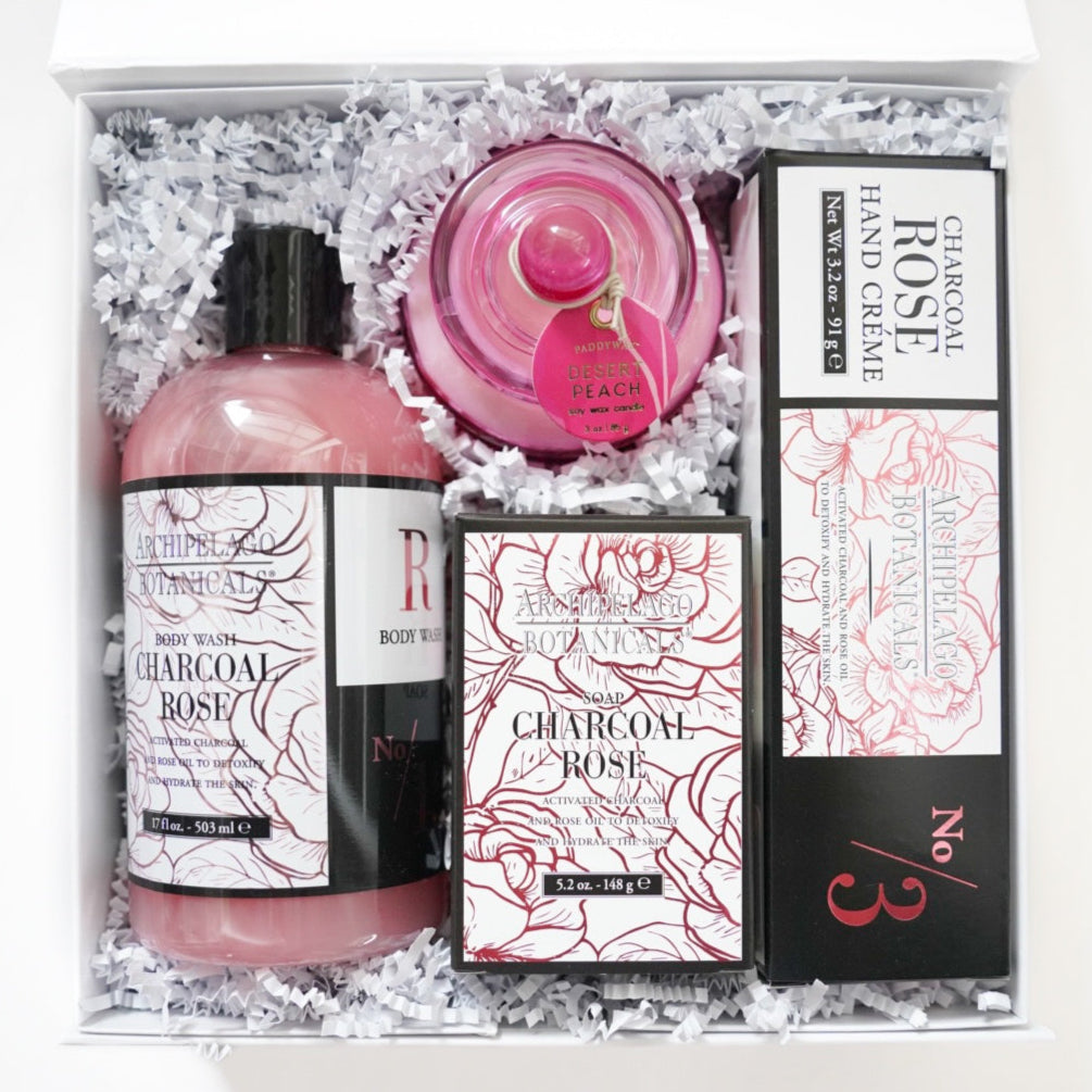Photograph of an Archipelago Botanicals gift set featuring Charcoal Rose bath products and a Paddywax Desert Peach candle nestled in white crinkle paper, showcasing the dusty pink liquids, intricate floral labels, and a vibrant pink glass jar lid
