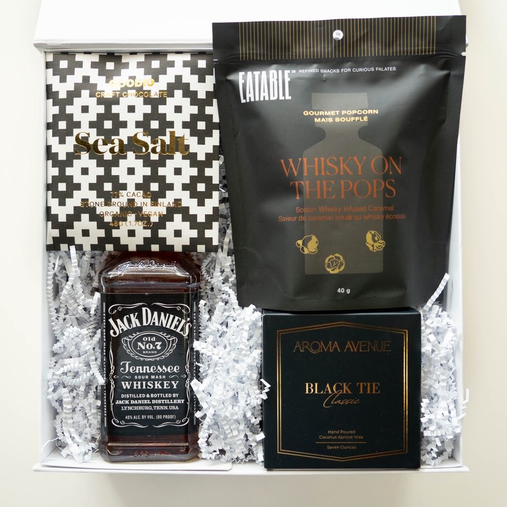 Photograph of a curated luxury gift box featuring a Jack Daniel's Tennessee Whiskey bottle, Eatable gourmet popcorn, and an Aroma Avenue candle nestled in white crinkle-cut paper with a black and white geometric pattern and gold-foiled text