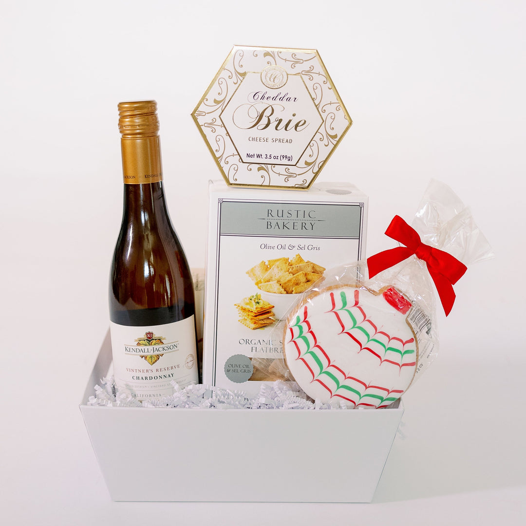 Photograph of a curated holiday gift box featuring Kendall-Jackson Chardonnay, Rustic Bakery flatbread, Cheddar Brie cheese spread, and a decorative red and green iced ornament cookie tied with a vibrant red bow