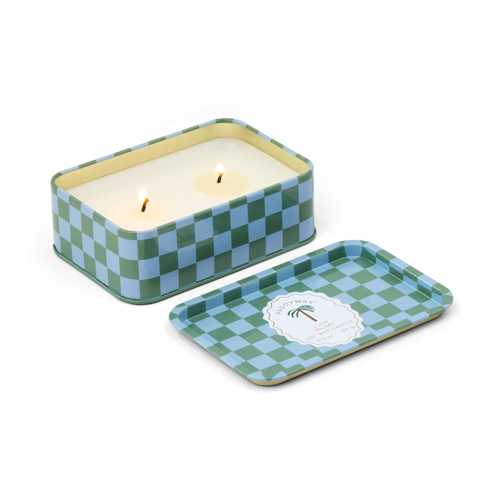 Soy wax candle by Paddywax features a rectangular Lush Palms scented tin on a white background with a green and blue checkered pattern, two lit wicks, and a scalloped label showing a palm tree symbol