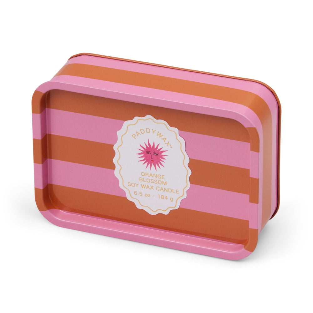 Metal tin and orange blossom soy wax candle by Paddywax featuring vibrant pink and orange horizontal stripes and a central scalloped label with a pink sun face illustration