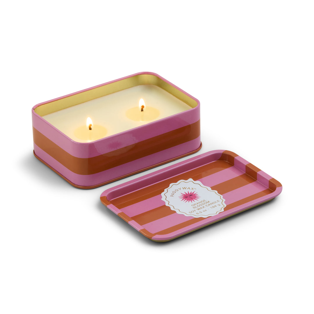 Product photograph by Paddywax of an Orange Blossom soy wax candle in a rectangular tin with pink and orange horizontal stripes featuring two glowing wicks, a gold-lined interior, and a matching lid with a sunburst logo on a white background