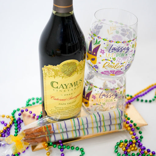 Bottle of Caymus Cabernet Sauvignon with a glass, Mardi Gras beads, and a wrapped gift on a white background