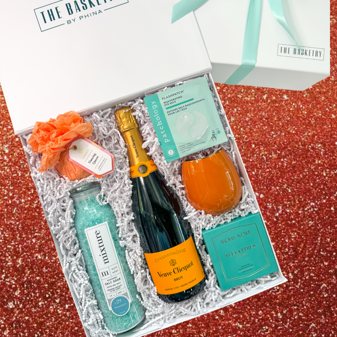 A product photograph by The Basketry by Phina of a luxury gift box featuring a bottle of Veuve Clicquot champagne, teal Mixture bath salts, and a vibrant orange tumbler nestled in white crinkle paper on a red glitter background