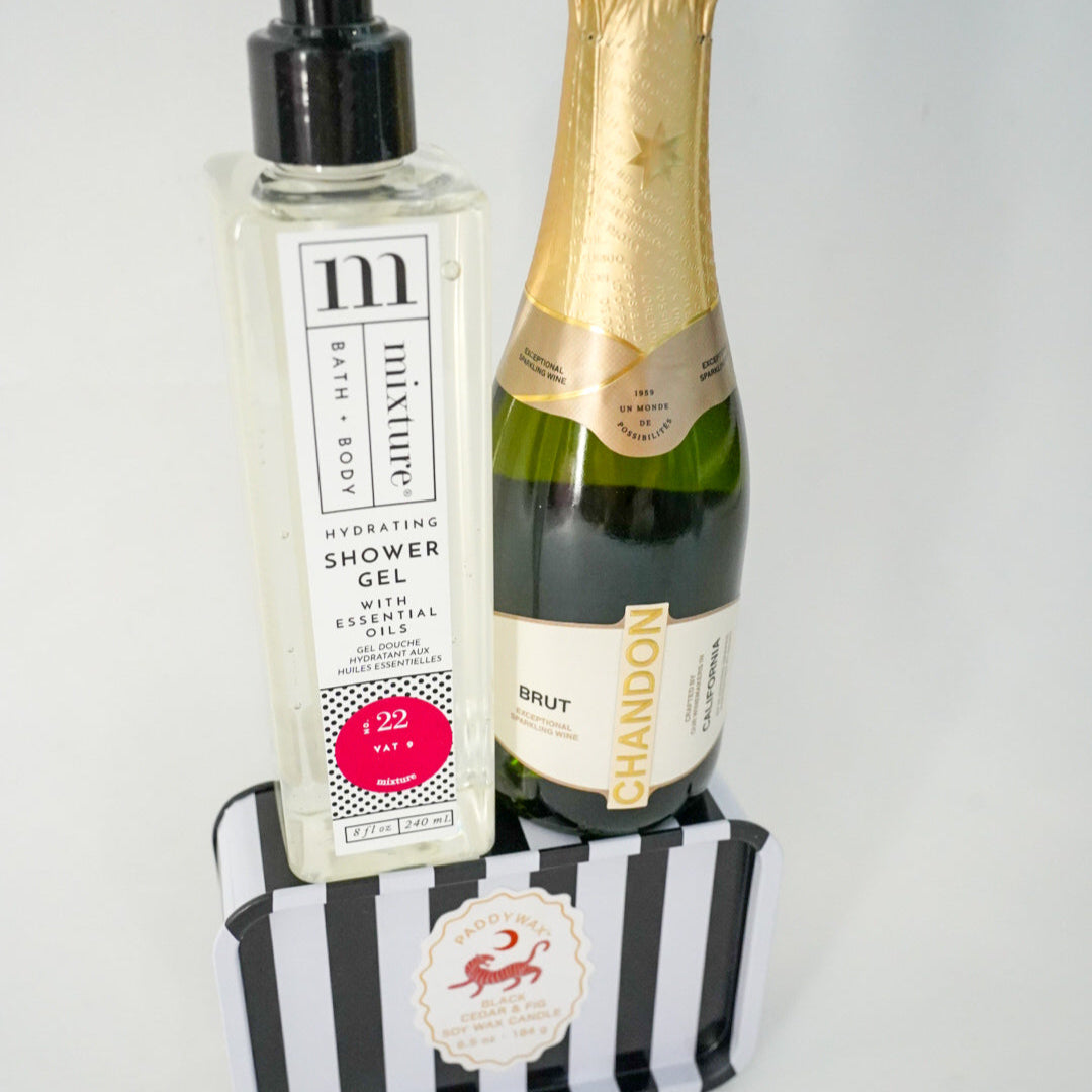 Digital photograph of a luxury gift arrangement featuring a bottle of Mixture Hydrating Shower Gel and Chandon Brut sparkling wine positioned on a black and white striped Paddywax soy wax candle tin, highlighting the vibrant hot pink circular label and metallic gold neck foil wrap against a clean white background