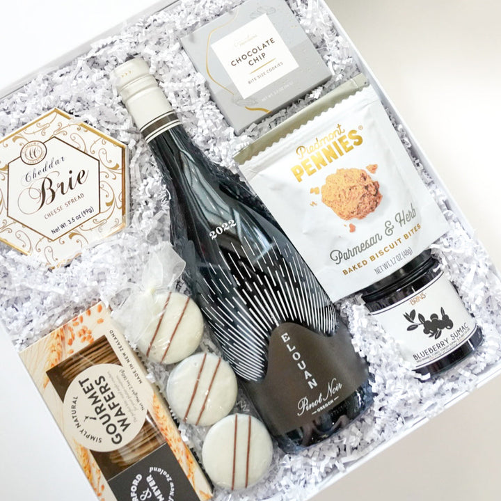 Photograph of a curated gourmet gift box featuring a 2022 Elouan Pinot Noir bottle, Piedmont Pennies parmesan biscuits, and blueberry sumac jam nestled in white crinkle paper with three striped white chocolate cookies and gold-accented brie cheese spread