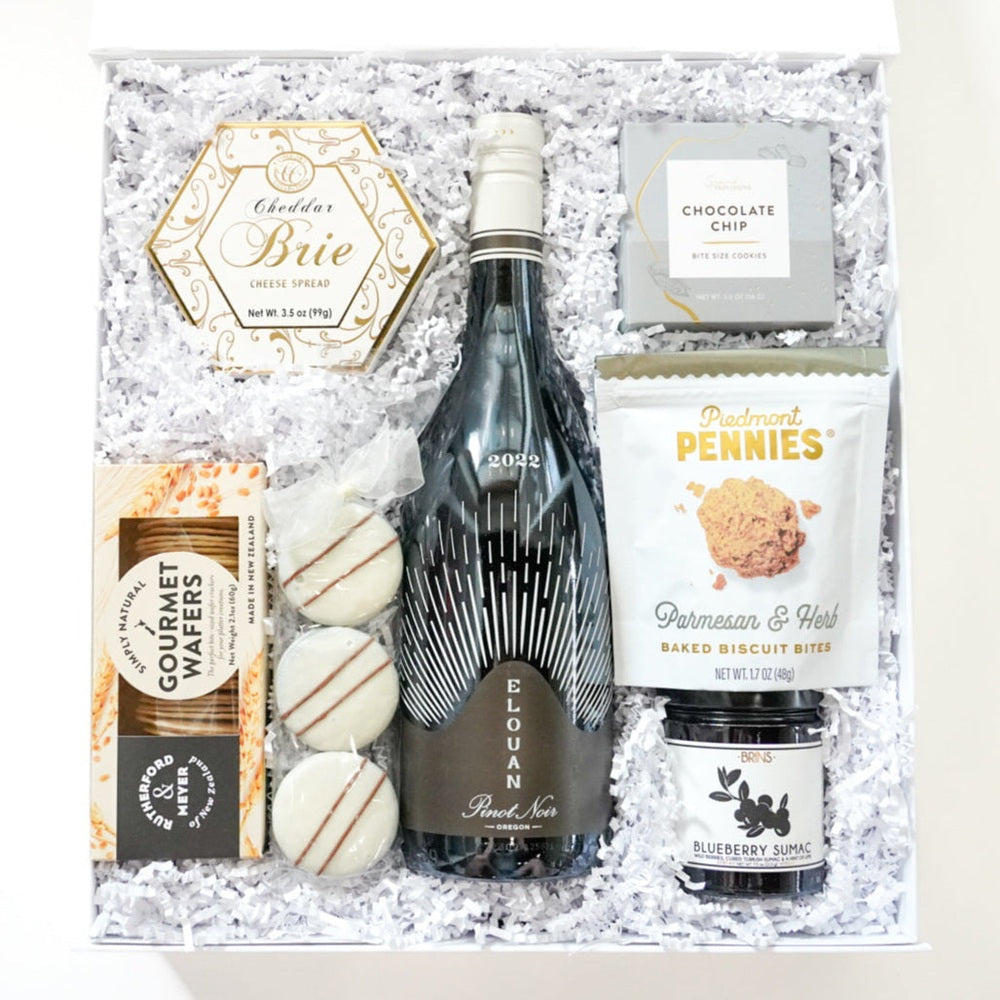 Photograph of a white gourmet gift box featuring a bottle of 2022 Elouan Pinot Noir from Oregon, a hexagonal gold-trimmed box of cheddar brie spread, and three white chocolate-drizzled cookies nestled in white shredded paper