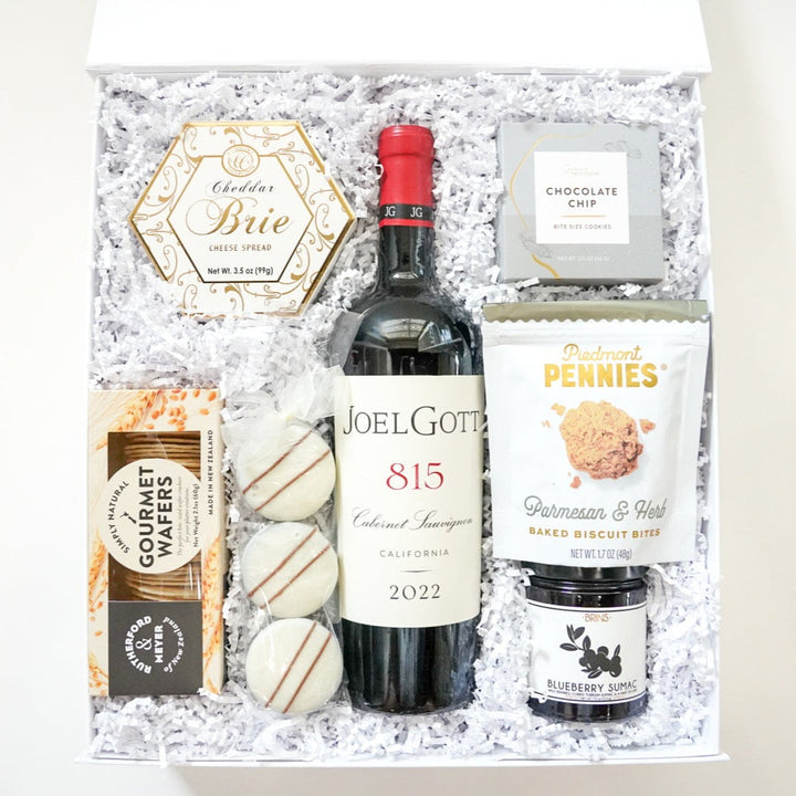 Photograph of a gourmet gift box containing a bottle of Joel Gott 815 Cabernet Sauvignon with its distinctive red cap, a gold-patterned hexagonal brie box, and three striped white chocolate treats on a bed of white crinkle paper