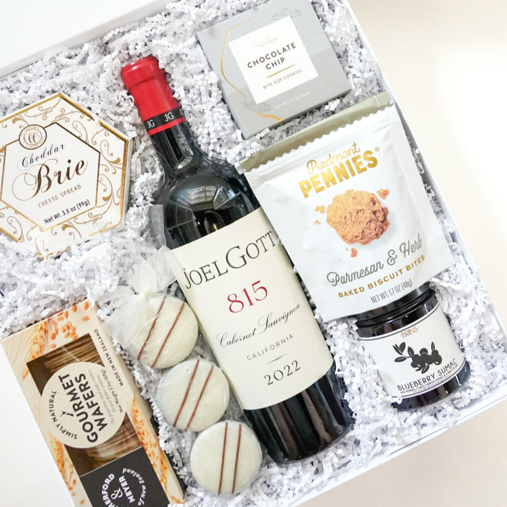 Photograph of a gourmet gift box featuring a bottle of 2022 Joel Gott 815 Cabernet Sauvignon, gold-labeled Cheddar Brie spread, and three striped white chocolate treats nestled in white crinkle paper with Piedmont Pennies snacks and Blueberry Sumac jam