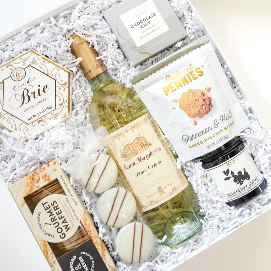 Photograph of a gourmet gift box containing a bottle of Santa Margherita Pinot Grigio, Piedmont Pennies biscuit bites, and three white chocolate-dipped cookies with brown stripes tied in a white ribbon and nestled in white shredded paper