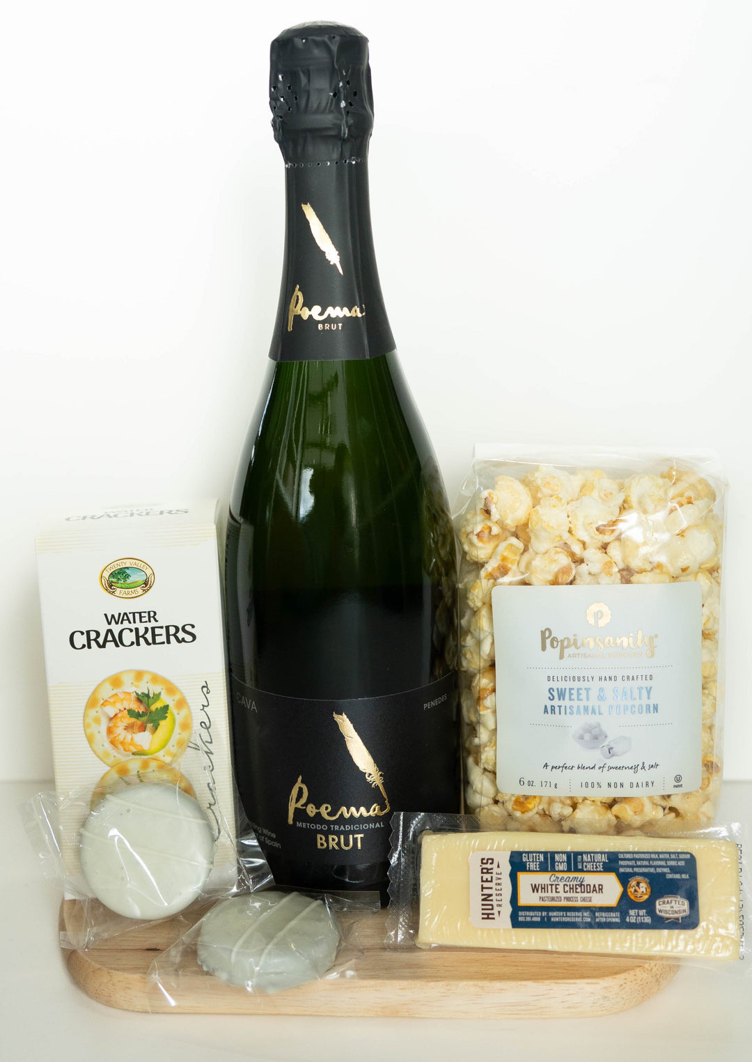 Photograph of a gourmet snack gift set on a wooden serving board featuring a bottle of Poema Brut Cava with a gold feather logo, a bag of Popinsanity artisanal popcorn, and two individually wrapped pale green confections against a white background