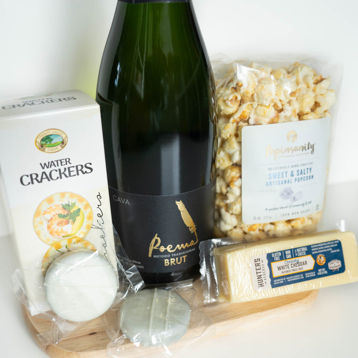 Photograph of a gourmet food gift set on a wooden cutting board featuring a dark green bottle of Poema Cava Brut with a gold feather logo, a bag of Popinsanity popcorn, a block of Hunter’s Reserve white cheddar cheese, and two round white-iced cookies in clear wrappers