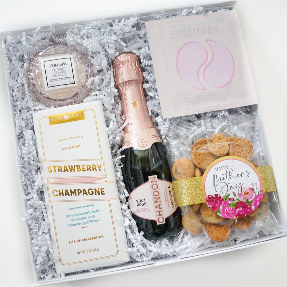 Photograph of a luxury Mother's Day gift box on a white background featuring a Chandon Brut Rosé bottle, Voluspa candle, and strawberry champagne chocolate bar nestled in white shredded paper with a gold glitter ribbon and pink floral accents