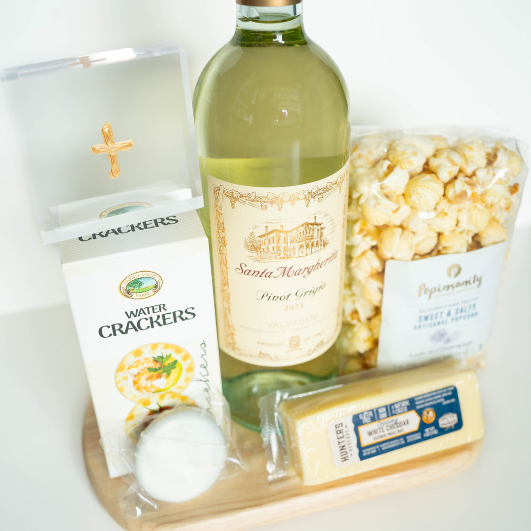 Professional studio photograph of a gourmet gift set on a wooden platter featuring a bottle of Santa Margherita Pinot Grigio with an illustrated villa on the label, Hunters white cheddar cheese, and a clear acrylic box with a gold cross