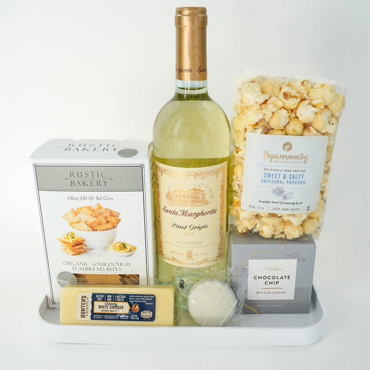 Studio photograph of a curated gourmet gift set featuring a bottle of golden Santa Margherita Pinot Grigio, textured Popinsanity artisanal popcorn, and Rustic Bakery sourdough flatbread bites arranged on a minimalist white tray