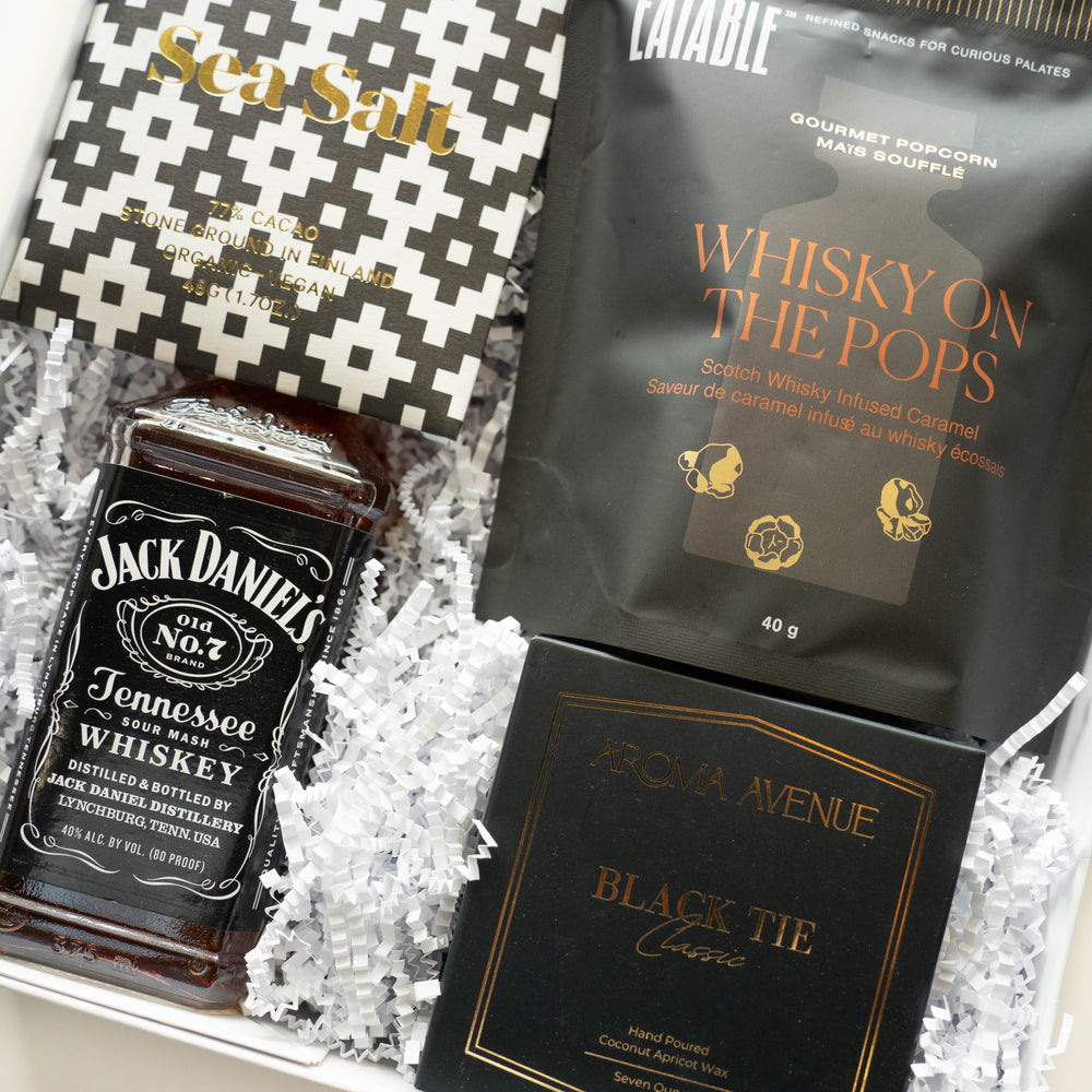 Photograph of a luxury curated gift box featuring an iconic bottle of Jack Daniel's Tennessee Whiskey, a bag of Eatable Whisky on the Pops popcorn, and a black and gold Aroma Avenue candle box nestled in white crinkle-cut paper filler with a geometric-patterned chocolate bar
