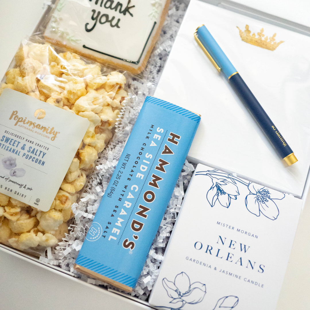 Commercial photograph of a curated thank you gift box featuring a New Orleans themed gardenia candle, a bright blue Hammond’s sea salt chocolate bar, and a white notepad with a gold crown crest and blue pen