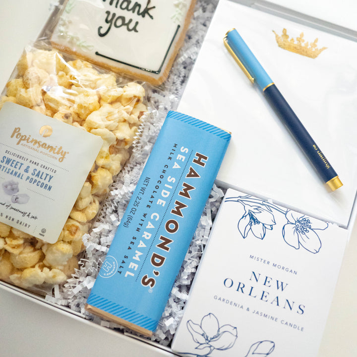 Commercial photograph of a curated thank you gift box featuring a New Orleans themed gardenia candle, a bright blue Hammond’s sea salt chocolate bar, and a white notepad with a gold crown crest and blue pen
