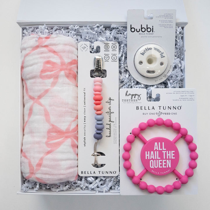 Product photograph by Bella Tunno of a baby gift set in a white box featuring a pink bow-patterned muslin swaddle, an ombre-shaded beaded pacifier clip, and a hot pink circular teether with the text All Hail The Queen