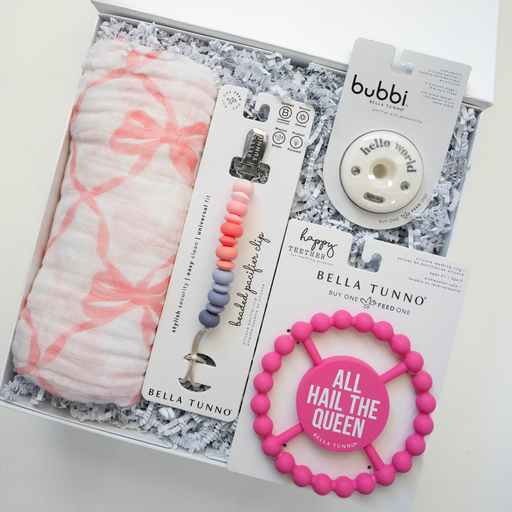 Photograph of Bella Tunno baby products curated in a white gift box featuring a pink bow-patterned muslin swaddle, an ombre beaded pacifier clip, and a bright pink teether with the text All Hail The Queen