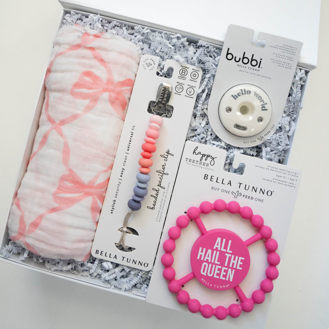 Photograph of Bella Tunno baby products curated in a white gift box featuring a pink bow-patterned muslin swaddle, an ombre beaded pacifier clip, and a bright pink teether with the text All Hail The Queen