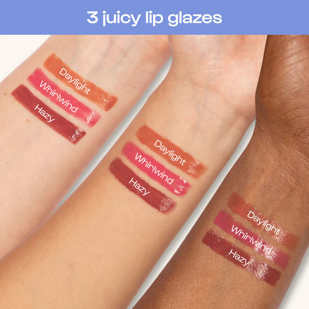 Commercial digital photograph of three juicy lip glaze swatches labeled Daylight, Whirlwind, and Hazy on inner forearms of three different skin tones featuring glossy peach, vibrant pink, and deep berry finishes