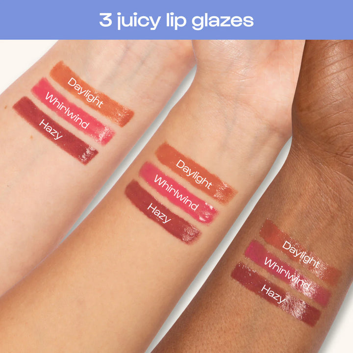 Commercial digital photograph of three juicy lip glaze swatches labeled Daylight, Whirlwind, and Hazy on inner forearms of three different skin tones featuring glossy peach, vibrant pink, and deep berry finishes