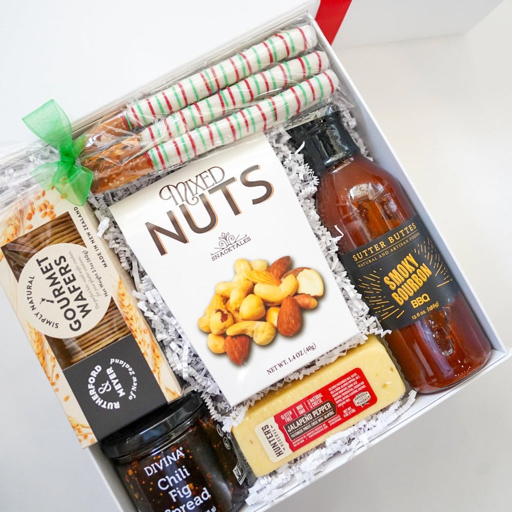 Photograph of a gourmet food gift box on a white background featuring a bottle of Sutter Buttes Smoky Bourbon BBQ sauce and Snacktales Mixed Nuts accented by festive red and green striped pretzel rods and a bright green ribbon bow
