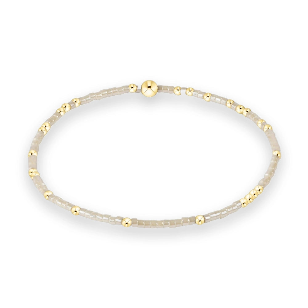 Beaded jewelry bracelet displayed on a white background featuring neutral cream-colored square beads, small polished gold spheres, and a prominent large central gold bead focal point