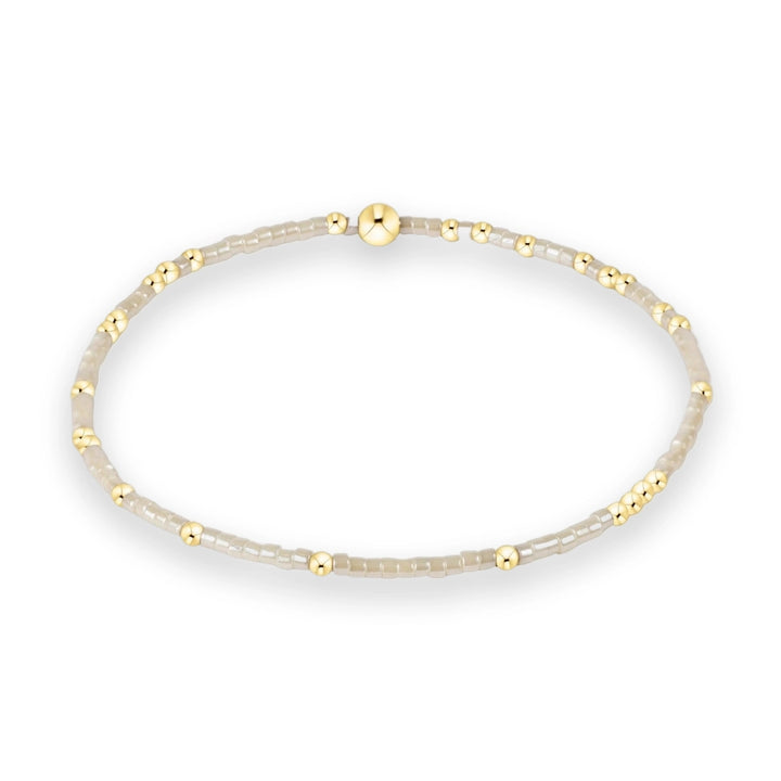 Beaded jewelry bracelet displayed on a white background featuring neutral cream-colored square beads, small polished gold spheres, and a prominent large central gold bead focal point