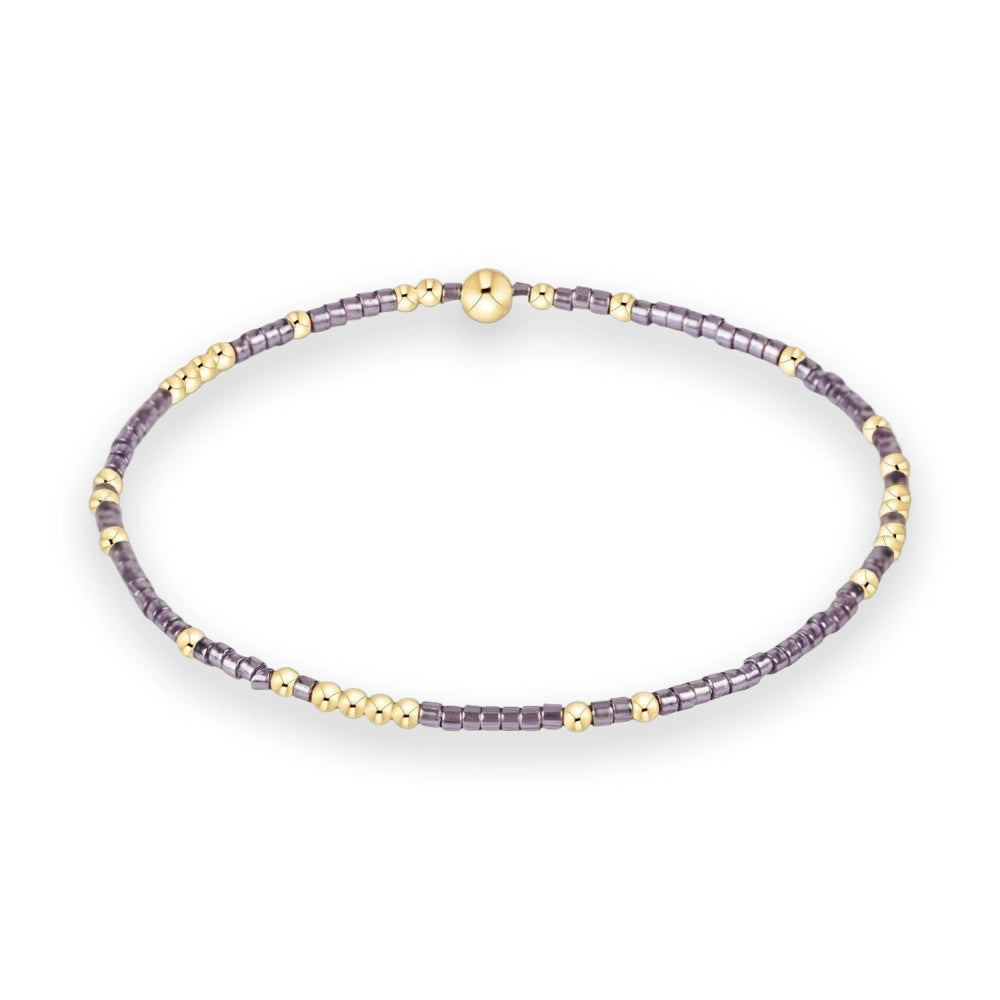 Beaded jewelry bracelet featuring iridescent purple cylinder beads and small polished gold spheres with a prominent large gold orb centerpiece on a minimalist white background
