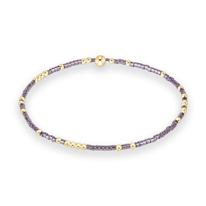 Beaded jewelry bracelet featuring iridescent purple cylinder beads and small polished gold spheres with a prominent large gold orb centerpiece on a minimalist white background