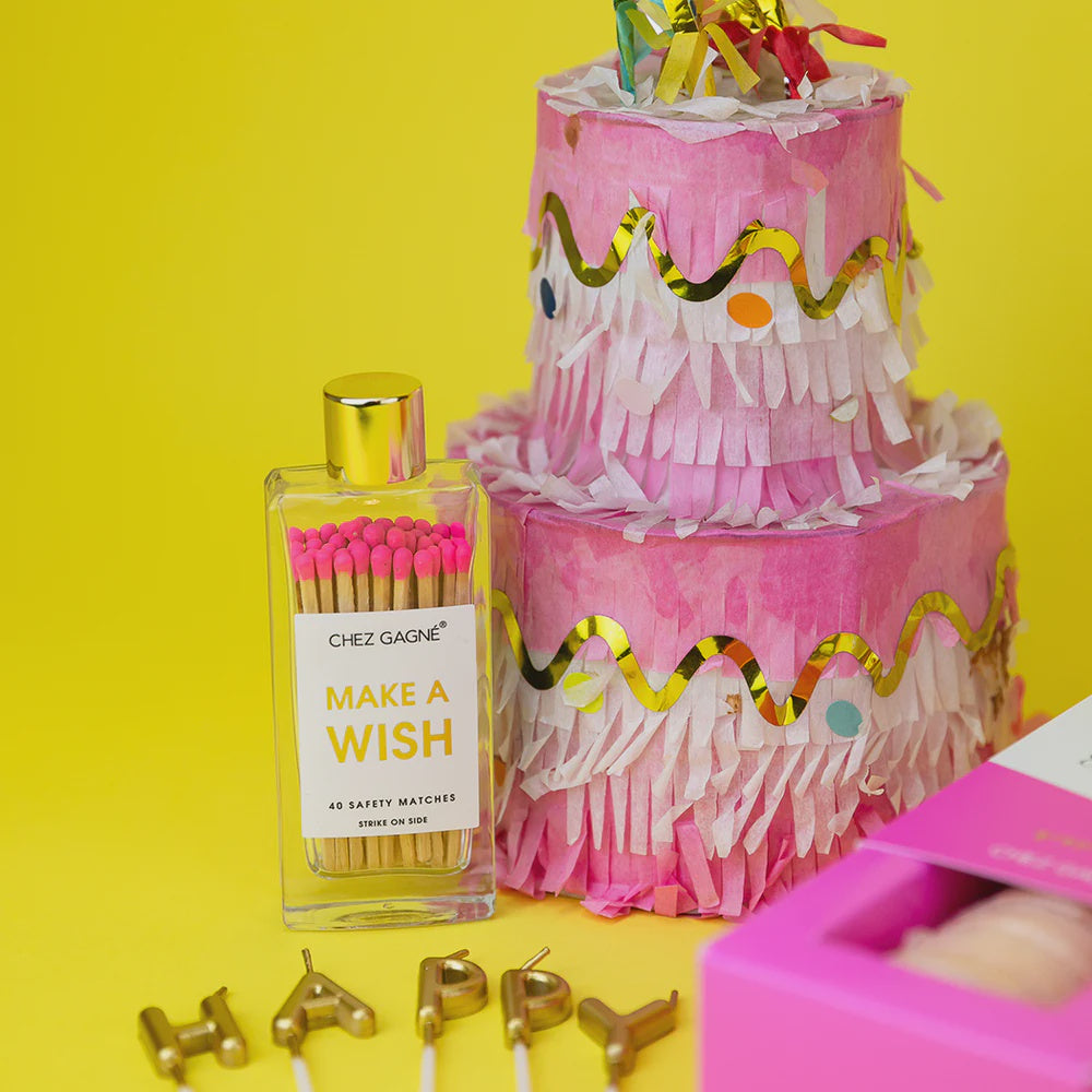 Commercial photograph by Chez Gagné of a Make A Wish glass bottle filled with pink-tipped matches and a pink tiered fringe piñata cake decorated with gold wavy trim and gold HAPPY candles on a yellow background