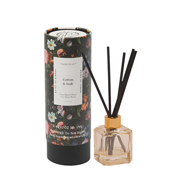 Studio photography by Paddywax showcasing a Cotton &amp; Teak glass reed diffuser set against a white background, featuring a dark cylindrical tube with vintage floral botanical illustrations and a square amber glass bottle with a metallic gold collar and four fanned-out black reeds