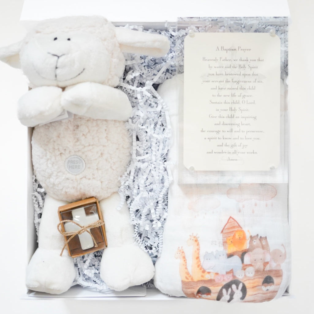Product photograph of a baptism gift set in a white box featuring a plush white lamb with a Press Here button, a small white cross tied with twine, and a muslin swaddle blanket illustrated with a colorful Noah's Ark including giraffes and lions