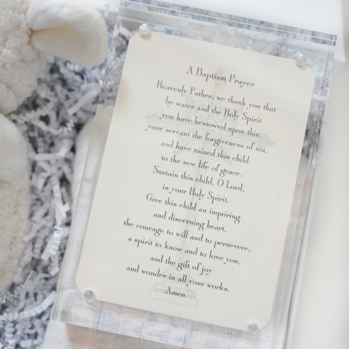 Photograph of a printed Baptism Prayer card inside a clear acrylic frame with silver bolts, featuring elegant serif typography and a subtle baptismal font illustration next to a soft white plush lamb in a gift box with shredded paper