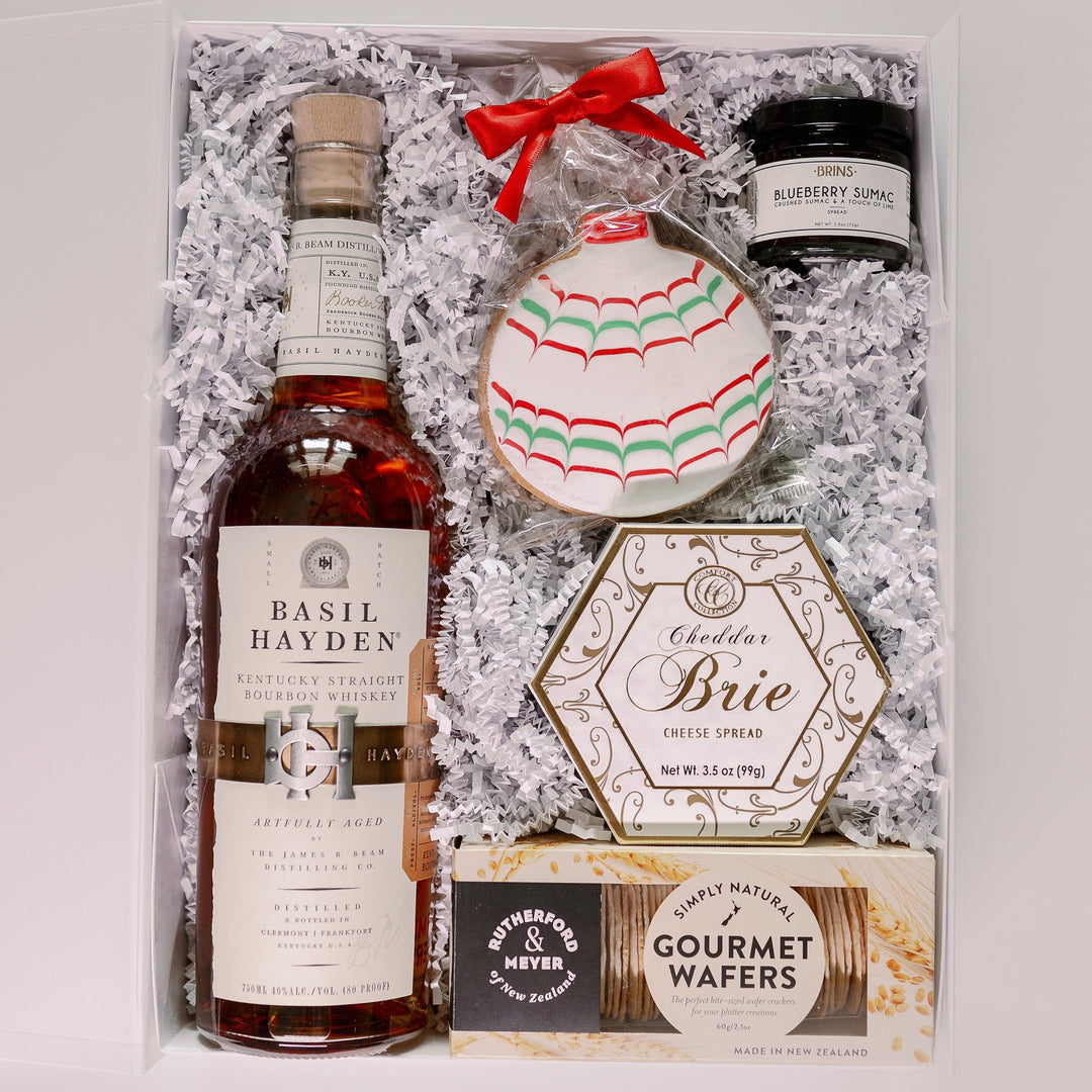 Digital product photograph of a gourmet holiday gift box featuring a bottle of Basil Hayden Bourbon and Rutherford & Meyer wafers nestled in white shredded paper alongside a hexagonal Cheddar Brie cheese spread and a round cookie decorated with red and green scalloped icing and a bright red bow