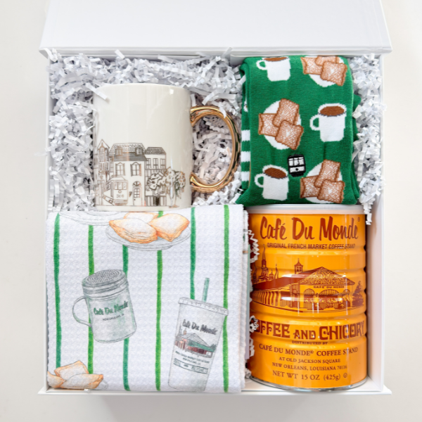 Photograph of a New Orleans souvenir gift box containing a gold-handled mug with streetcar illustrations, green beignet-print socks, and a bright orange Café Du Monde coffee tin featuring the St. Louis Cathedral landmark