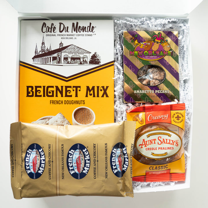 Digital photograph of a New Orleans gift box showcasing regional delicacies including a bright yellow Café Du Monde beignet mix box featuring the St. Louis Cathedral and French Market landmarks, purple and gold striped amaretto pecans, and a metallic gold bag of coffee
